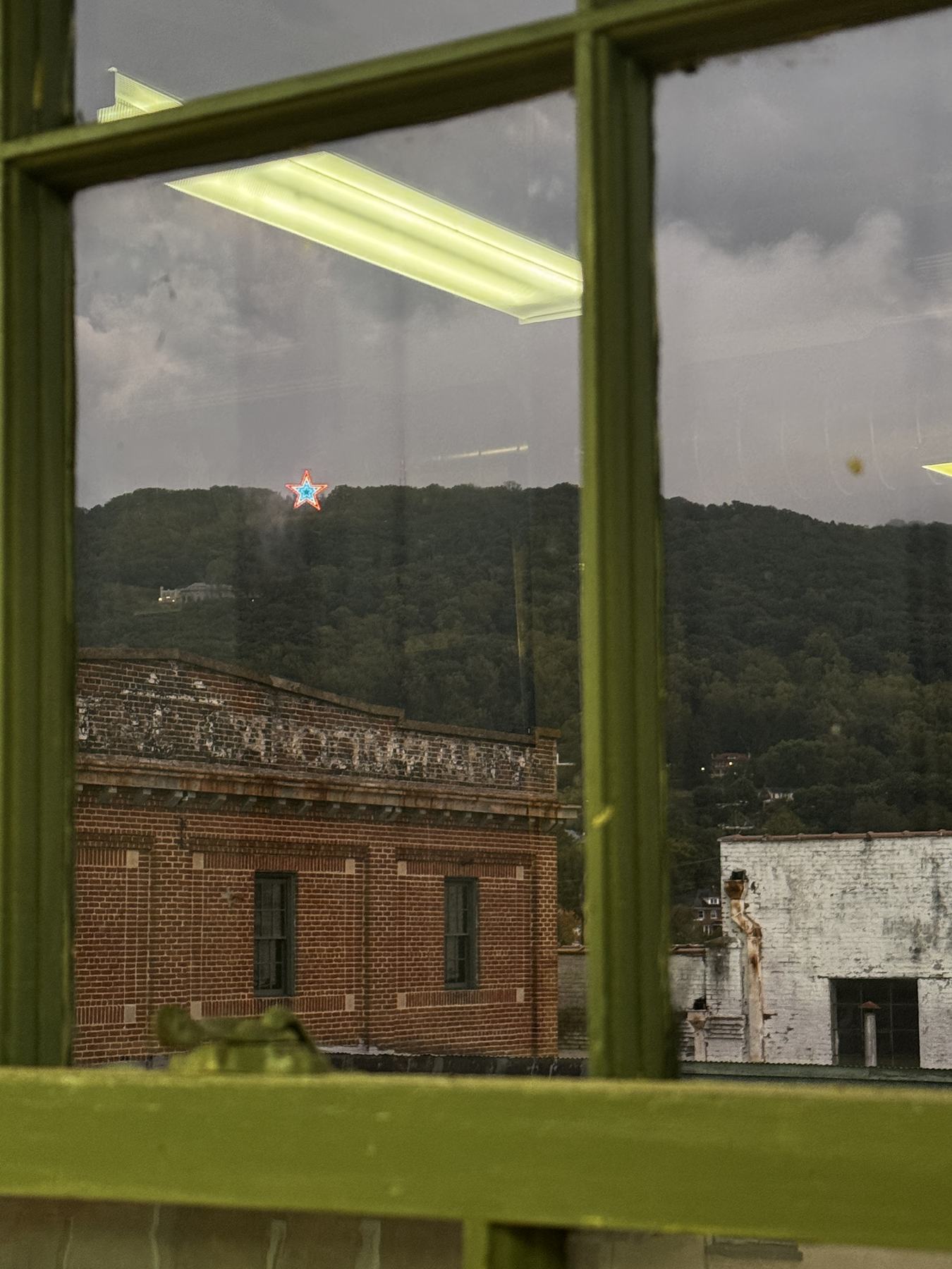 The Mill Mountain Star lit on the hillside at dusk, seen through a green-painted window sash above the brick parapet of the south building.